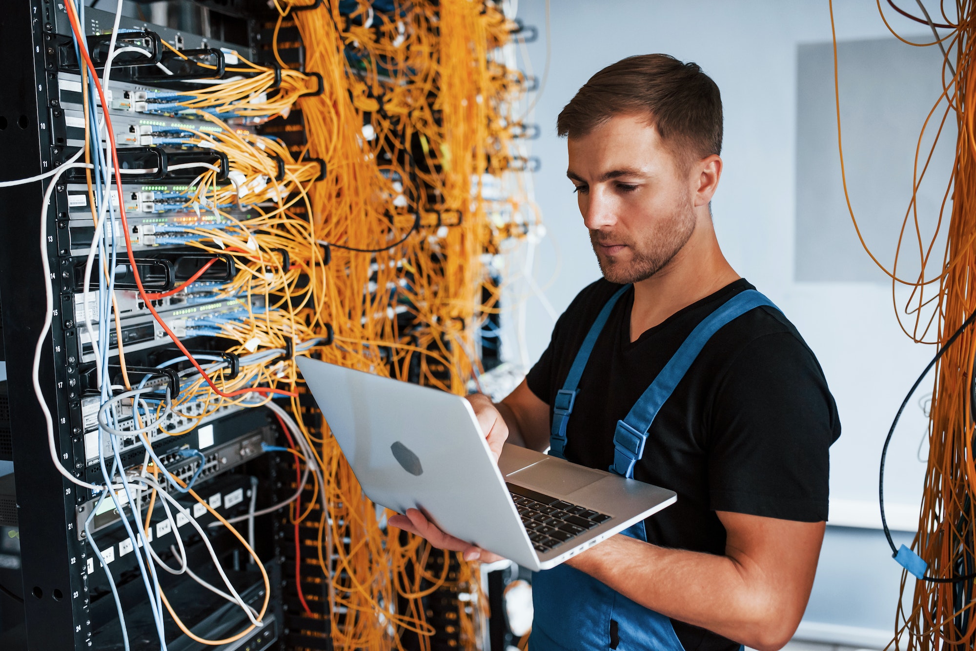 Young man in uniform and with laptop works with internet equipment and wires in server room